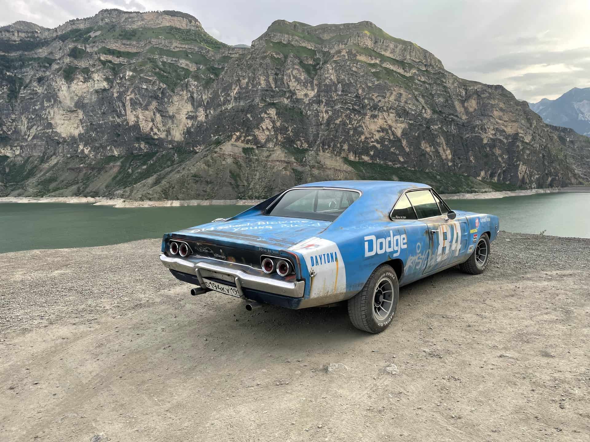 A classic blue Dodge Charger with racing livery parked beside a mountain lake