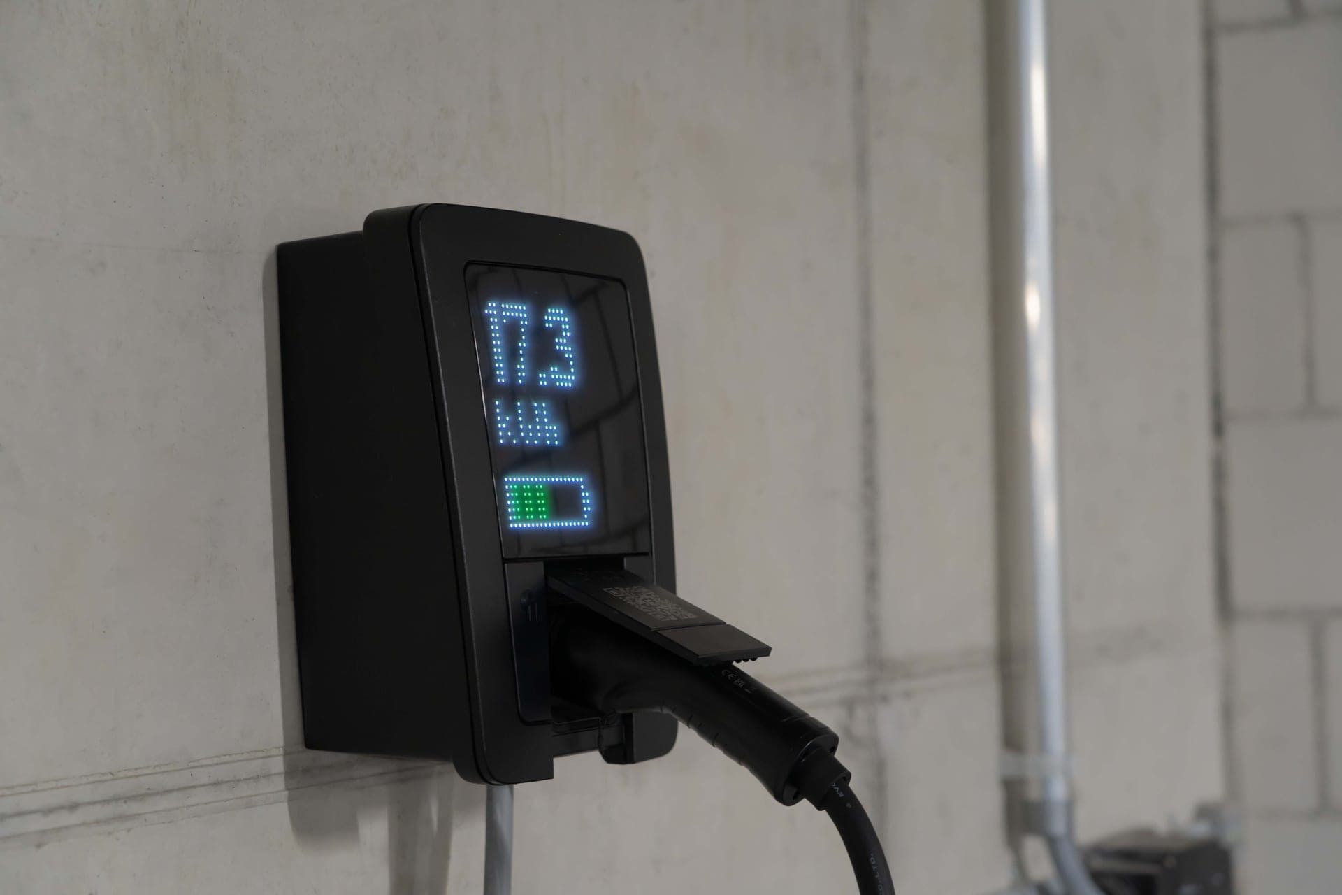 A black wall-mounted EV charger with a small display, its cable plugged in