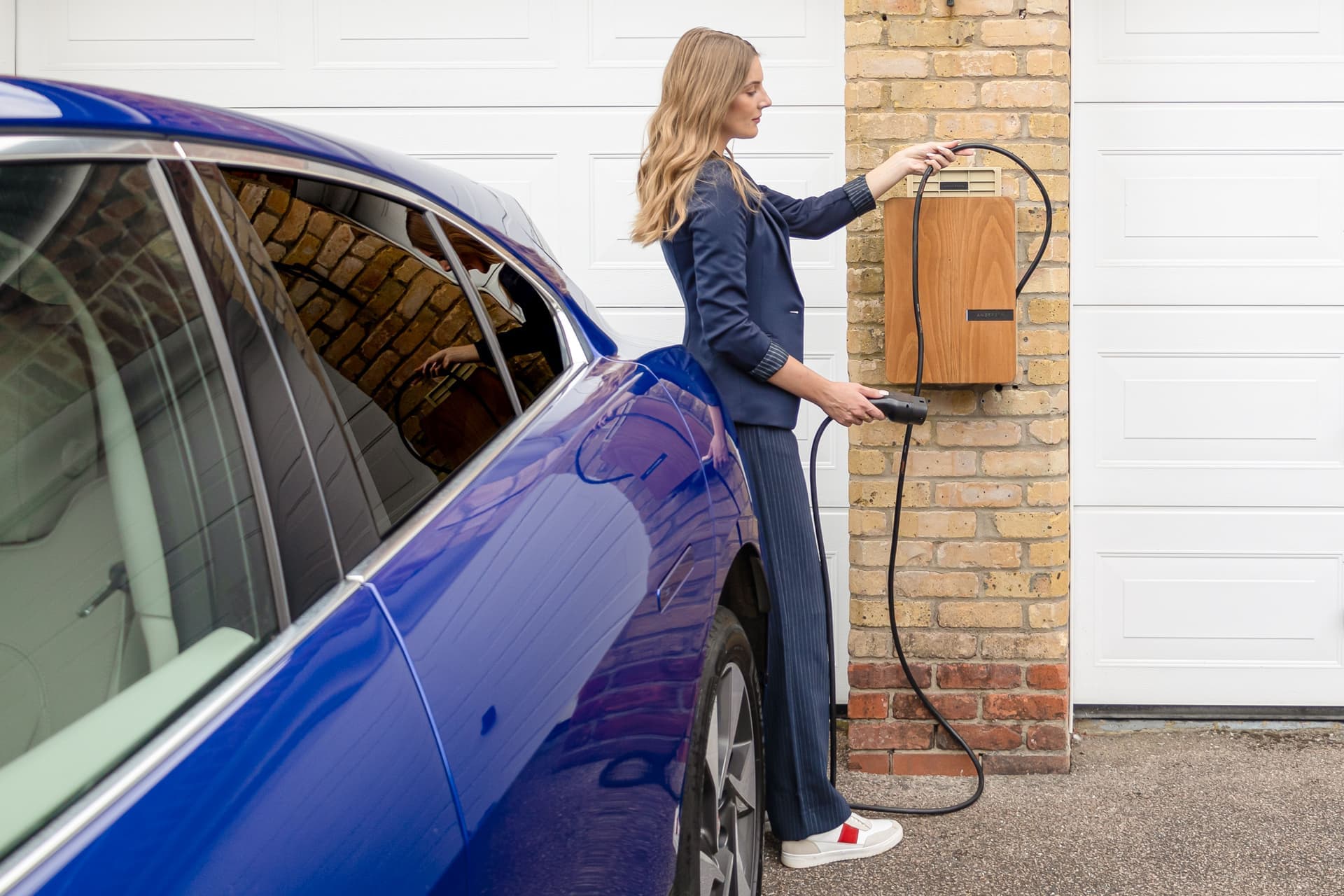 A woman lifting a charging connector from a wood-paneled wall charger beside a blue car