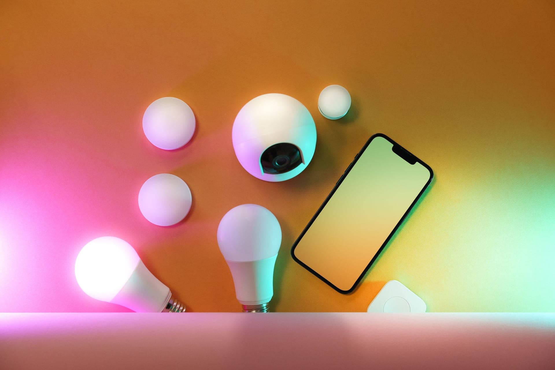 A smartphone beside smart bulbs, sensors and a home camera on a table