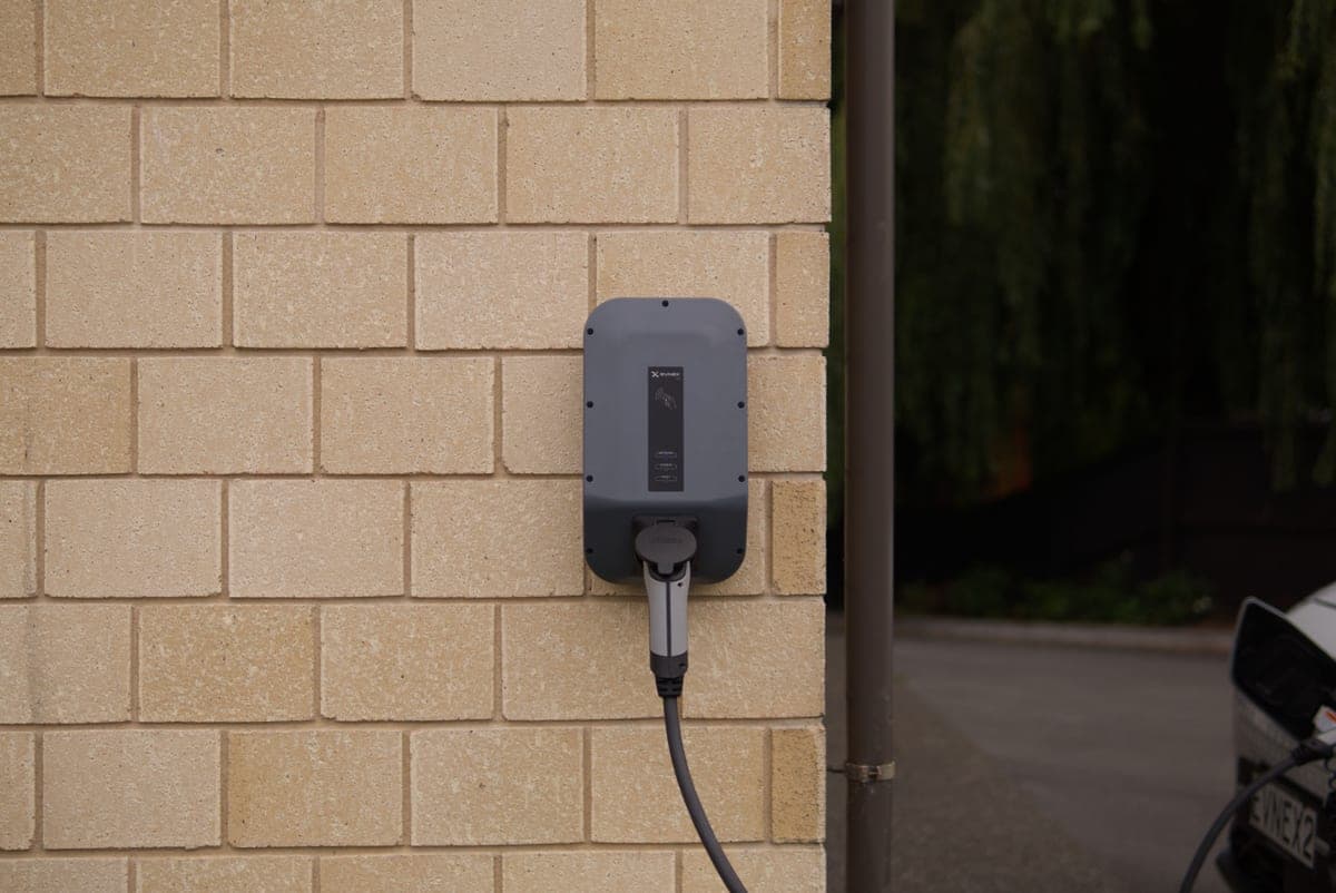A gray wall-mounted EV charger on a brick wall, its cable running to a parked car
